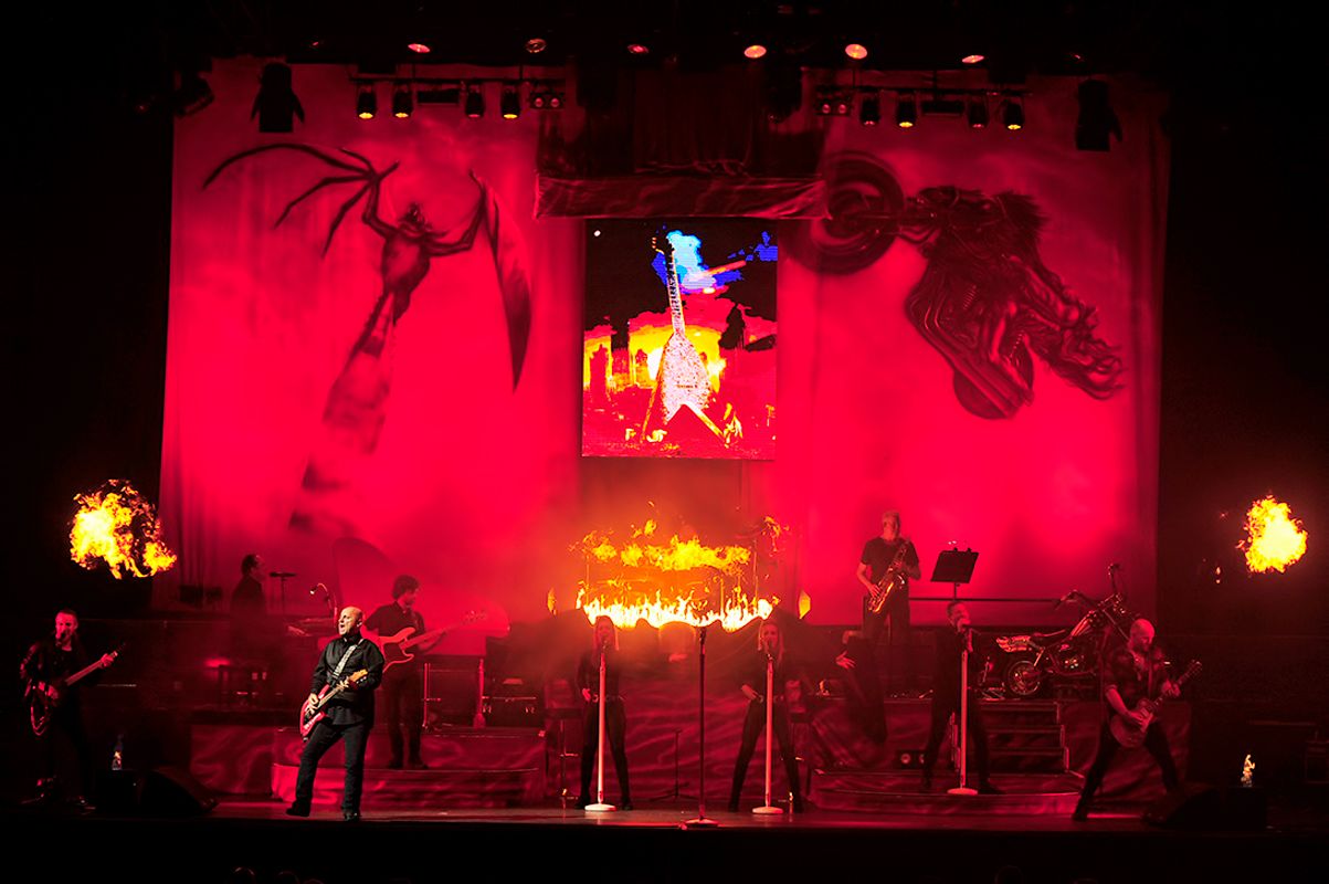 Steve Steinman and band performs on stage with an intense red backdrop featuring abstract designs. Flames burst from the stage, creating a dramatic and energetic atmosphere.