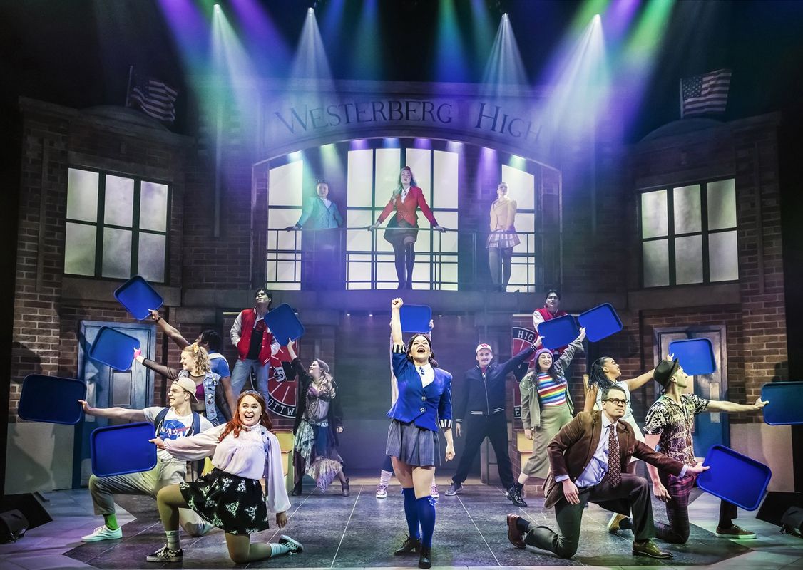 A vibrant stage scene shows performers in school uniforms celebrating under colorful lights. The backdrop reads "Westerberg High." Some hold blue trays aloft energetically.