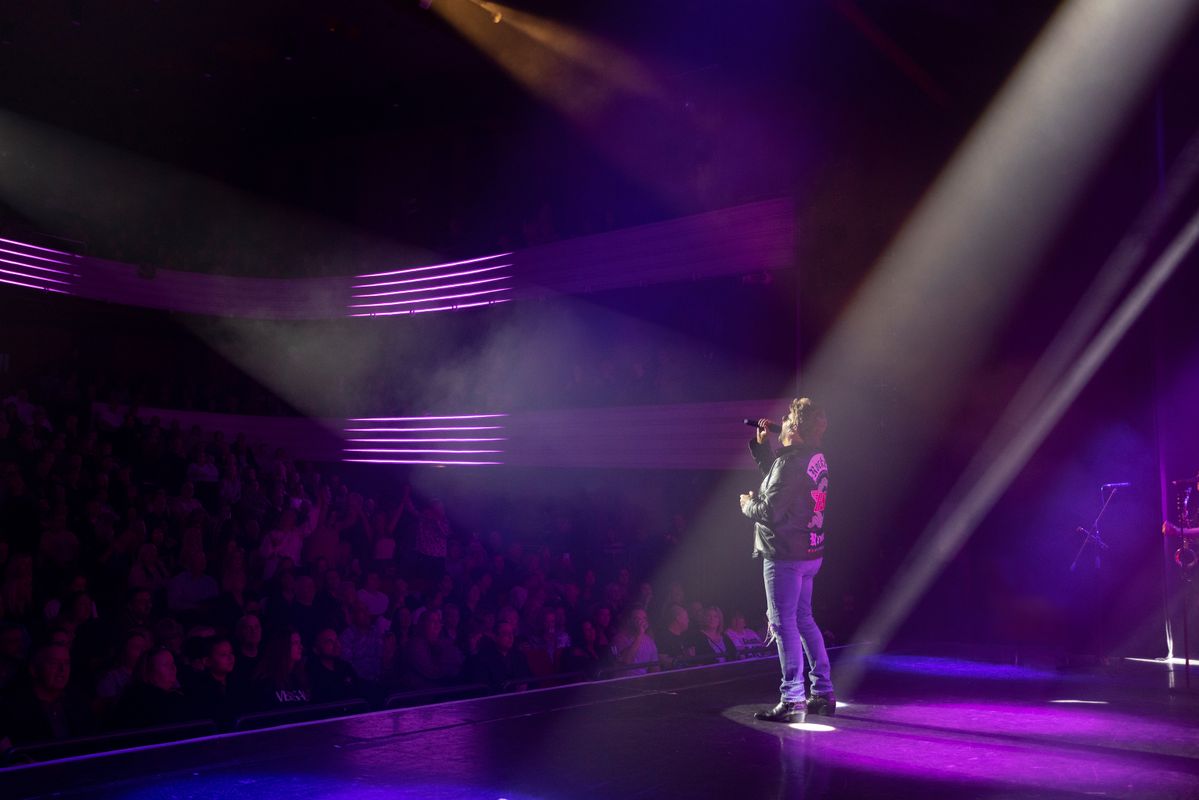 A man performing on stage to an audience. There are colourful lights. Behind him are backup singers and a band playing