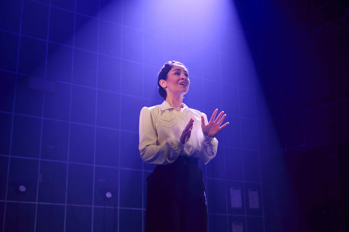 An actor in a white shirt performs on stage under dramatic blue lighting, with an expressive, intense gesture and facial expression, against a grid-patterned backdrop.