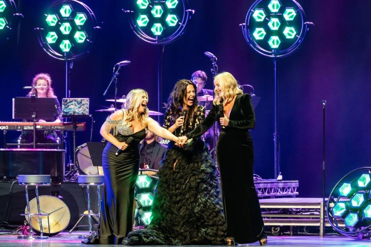 Kerry Ellis, Louise Dearman and Rachel Tucker in elegant gowns sing and laugh on stage, holding hands. Bright green lights and musicians in the background create a lively atmosphere.
