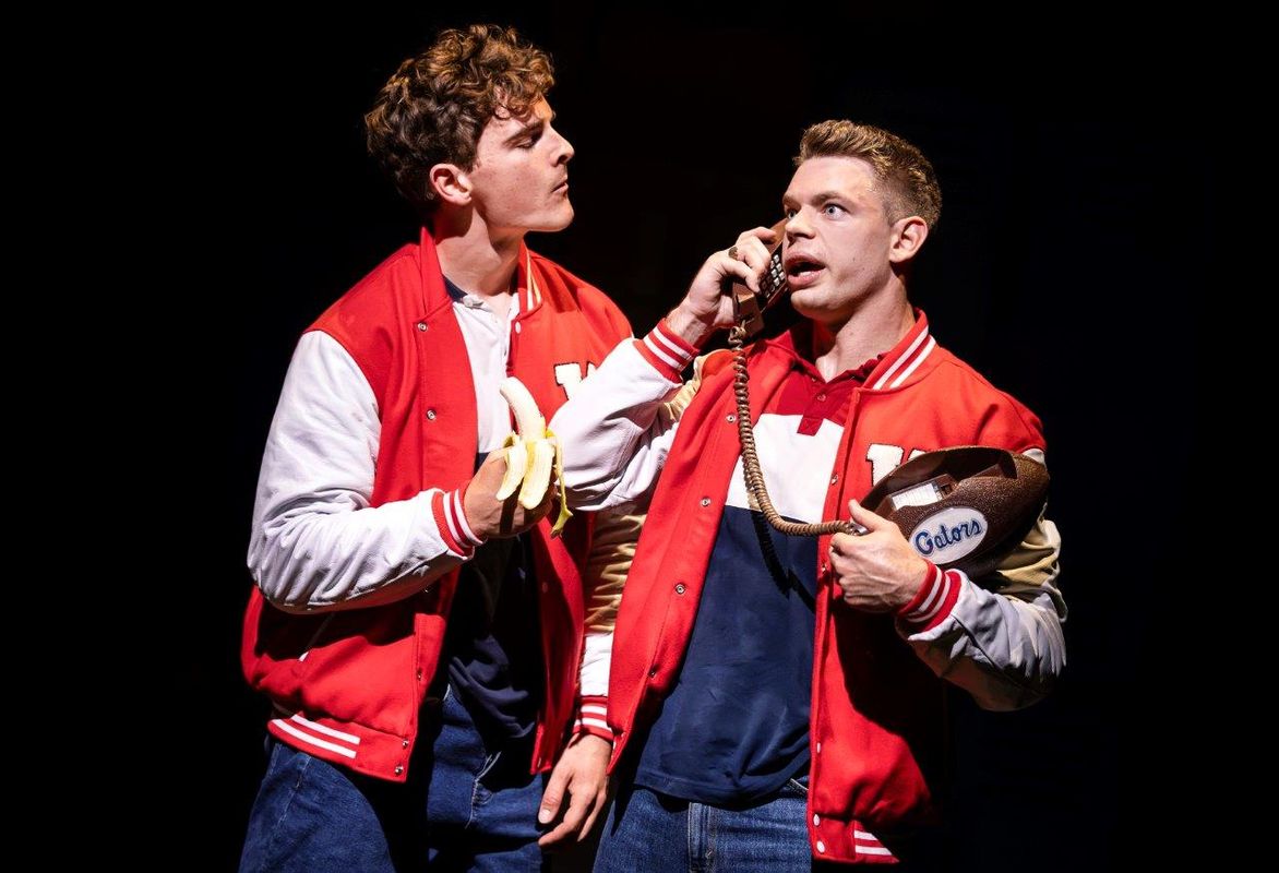 Two men in red and white jackets, one is eating a banana and the other is holding a football and talking on the phone