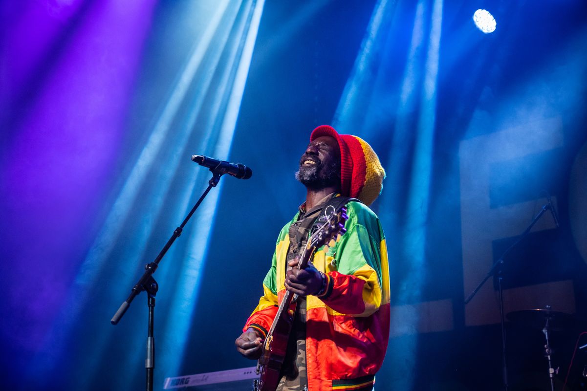 Musician performing passionately on stage, wearing a vibrant Rastafarian jacket and hat. Purple and blue lights create a lively, energetic atmosphere.