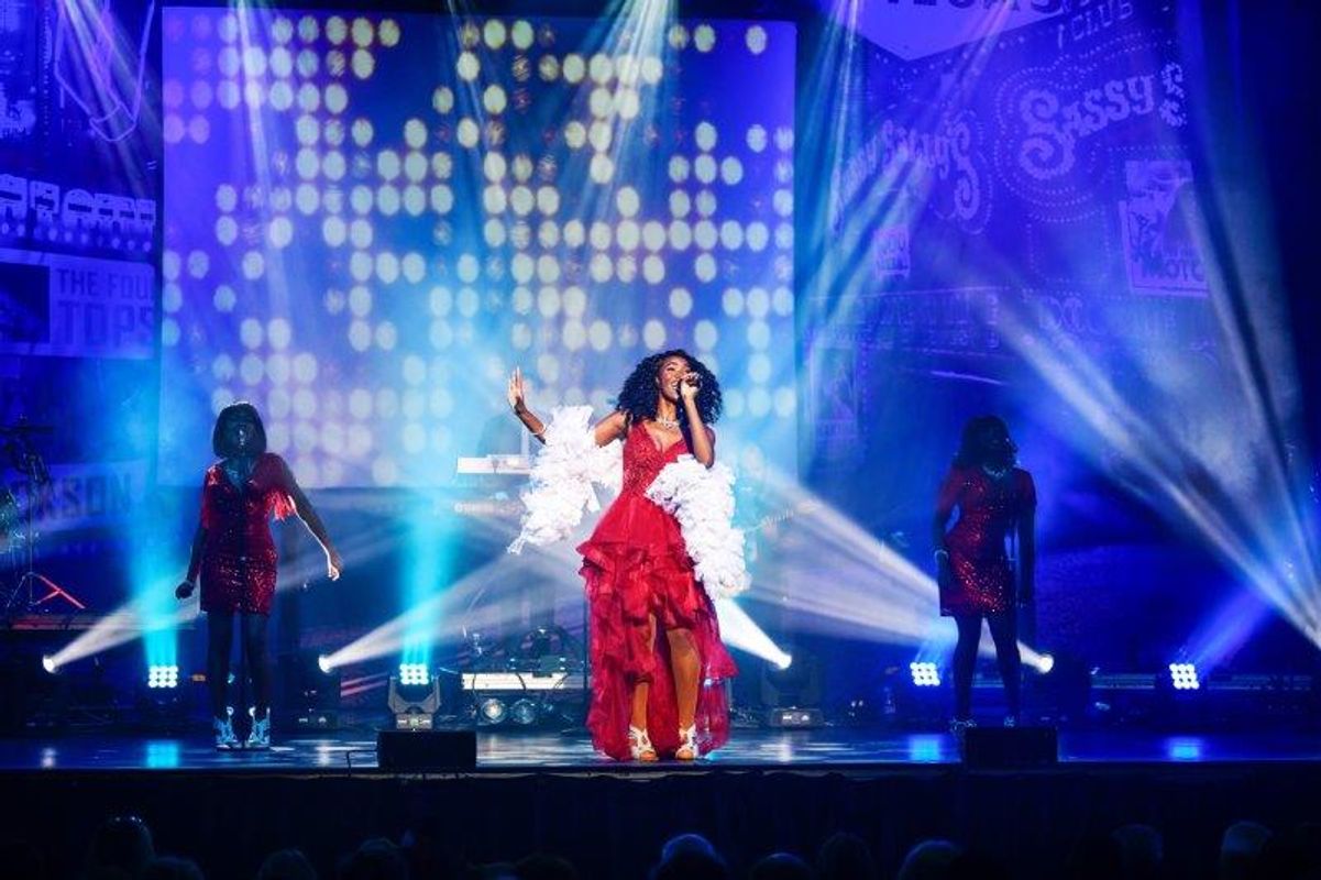 A person is stood centre stage wearing a red dress and white shawl.