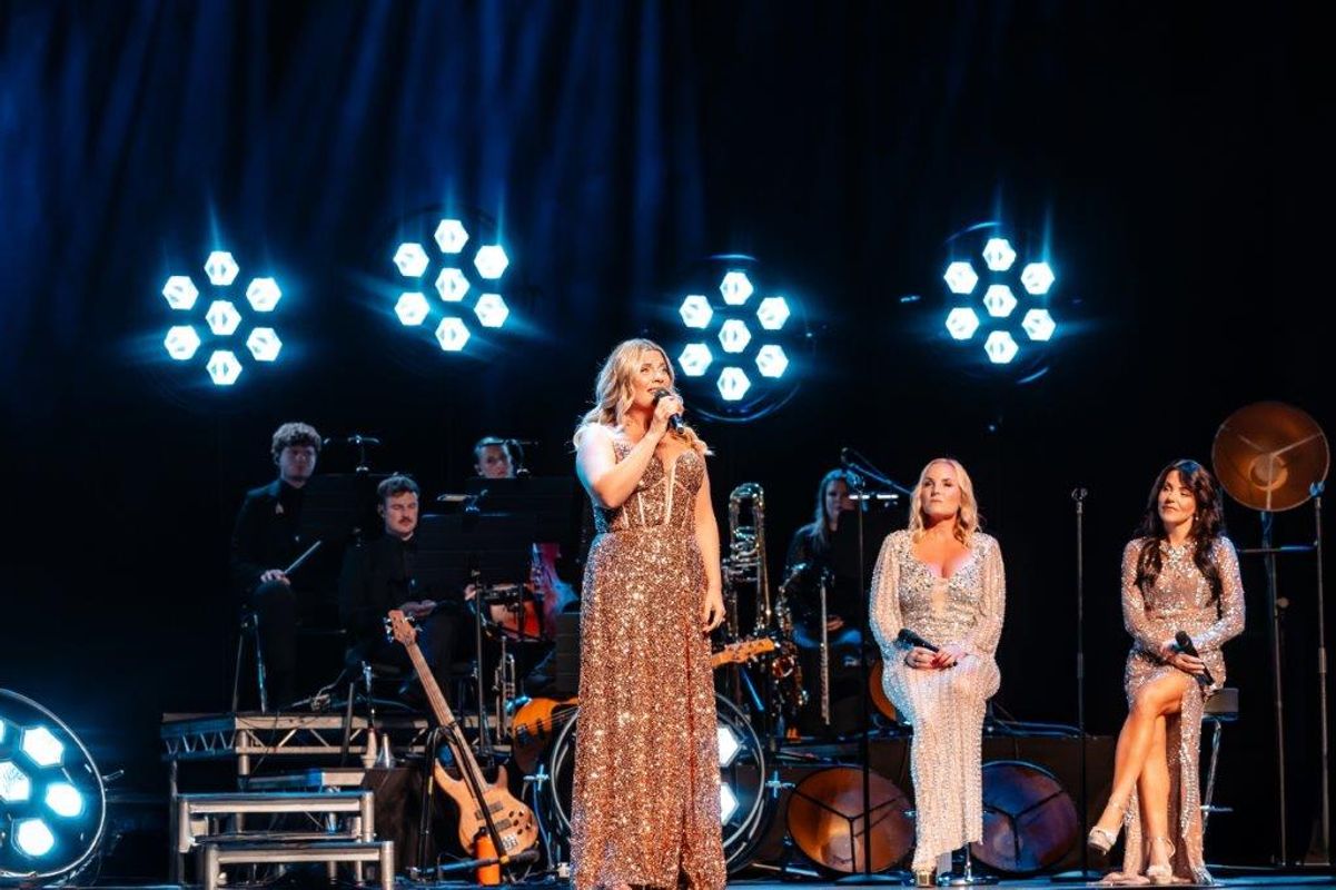 A singer in a glittering gown performs on stage with a live band. Two seated women in sparkling dresses watch, with bright spotlights behind them.