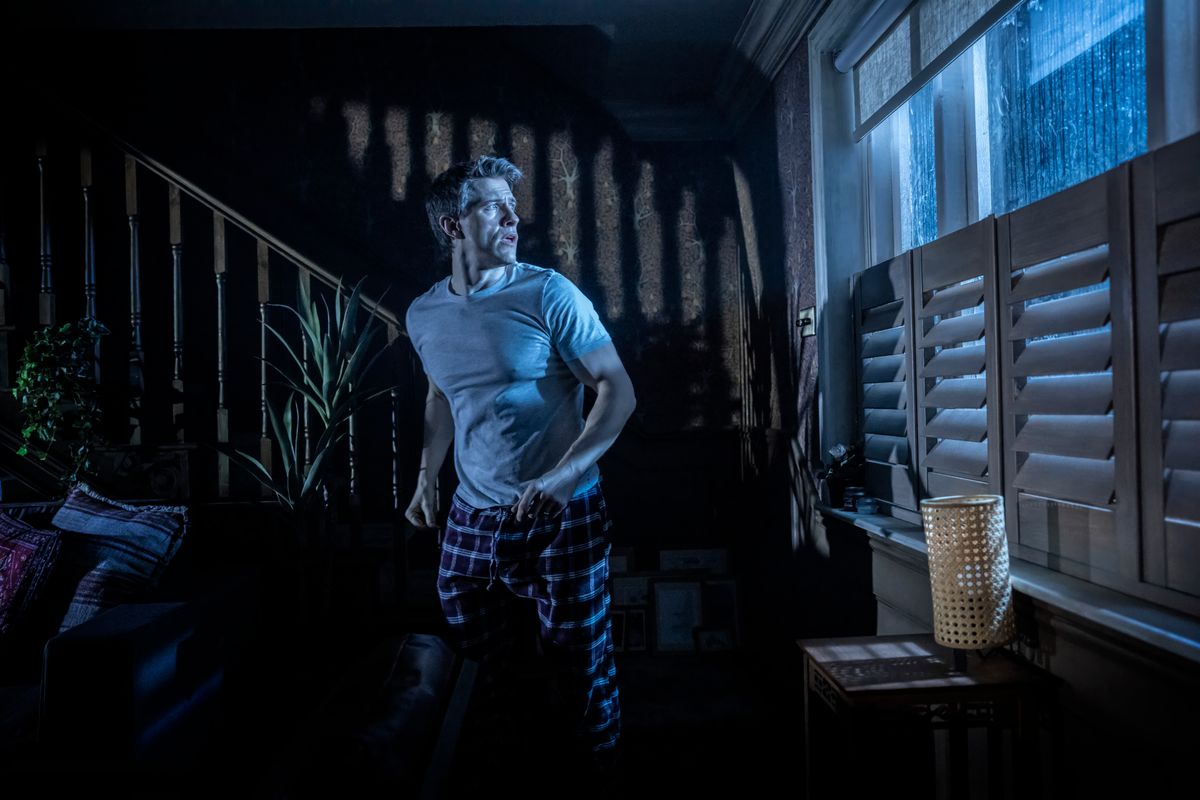 A man in pajamas anxiously looks out a window in a dimly lit room during a storm. Shadows from a staircase add drama, enhancing a tense atmosphere.