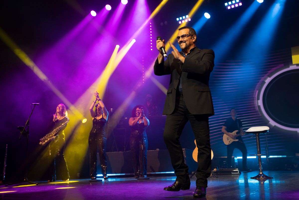 A man performing on stage to an audience. There are colourful lights. Behind him are backup singers and a band playing