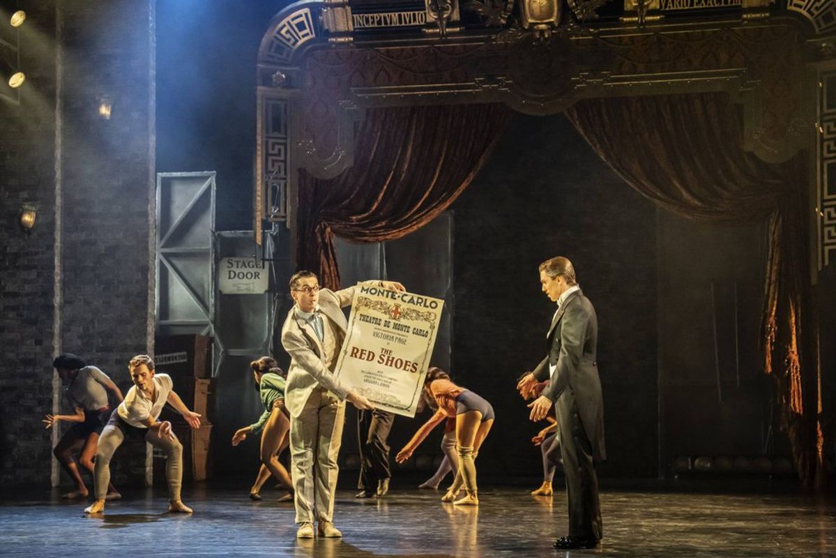 A person stands on stage holding a show advertisement for The Red Shoes.