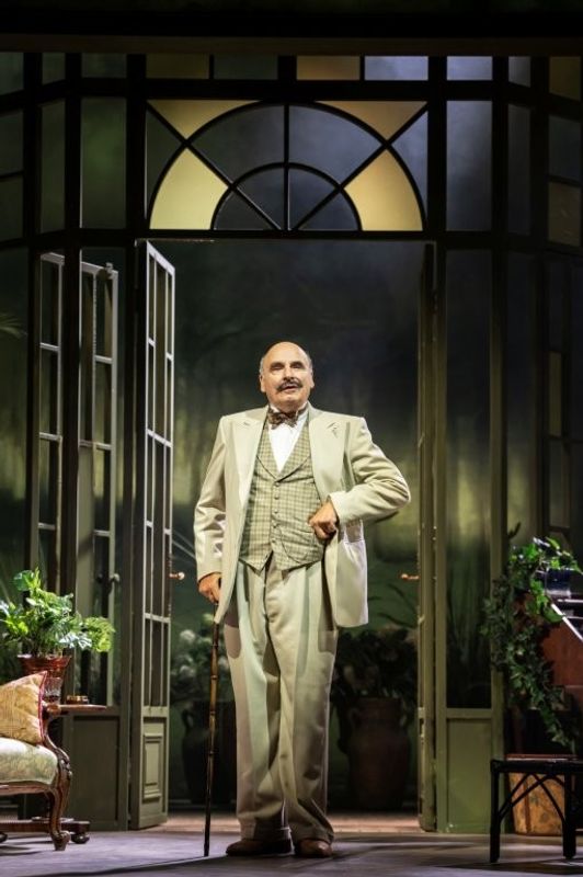A person in a formal, light-colored suit stands confidently in front of the open doors of a grand entrance. Behind them, lush greenery and potted plants are visible. The setting appears elegant and theatrical, with warm lighting casting soft shadows.