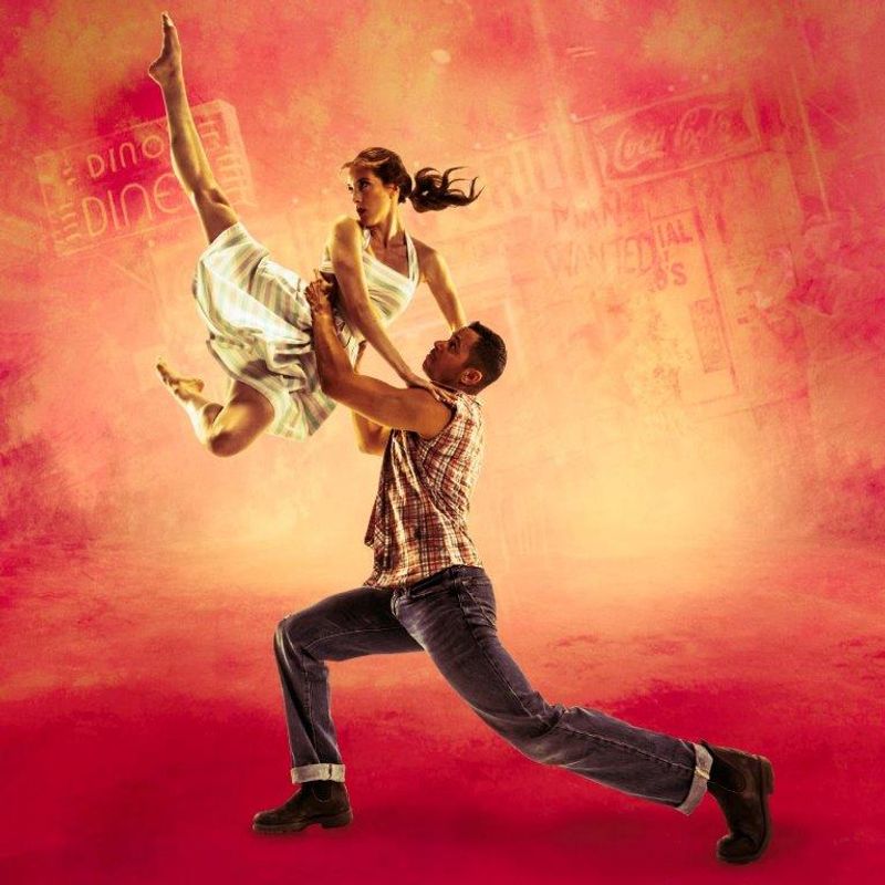 A male dancer in jeans lifts a female dancer in a white dress against a vintage diner backdrop. The vibrant pink and orange tones convey energy.