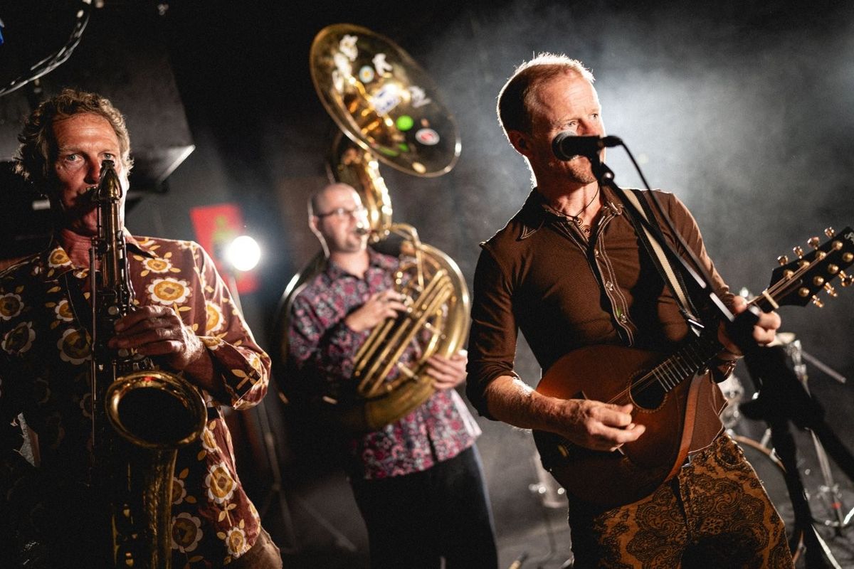 Three musicians perform energetically on stage. One plays a saxophone, another a sousaphone, and the third sings while playing a string instrument. The setting has dim lighting with a spotlight effect, creating a lively atmosphere.