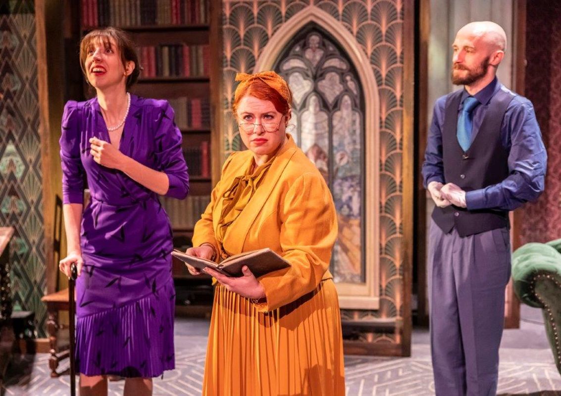 Three actors performing in a play, one in a purple dress with a cane, another in an orange outfit holding a book, and a third in a vest and tie. The setting resembles an elegant library with bookshelves and decorative arch. The mood is dramatic and intense.