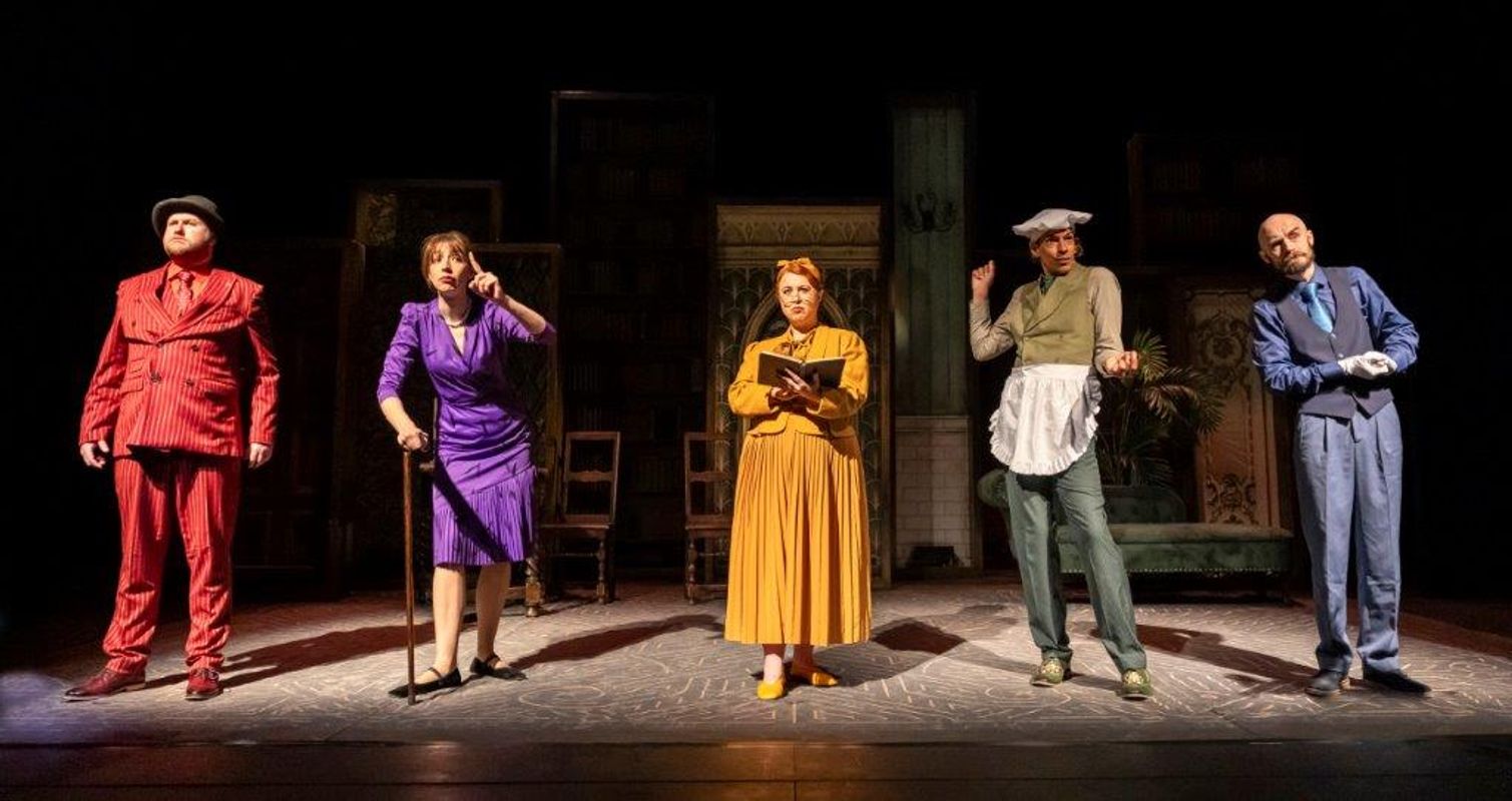 Five actors in vibrant costumes stand on stage, each striking a distinct pose. The mood is theatrical and lively, set against an eclectic backdrop.