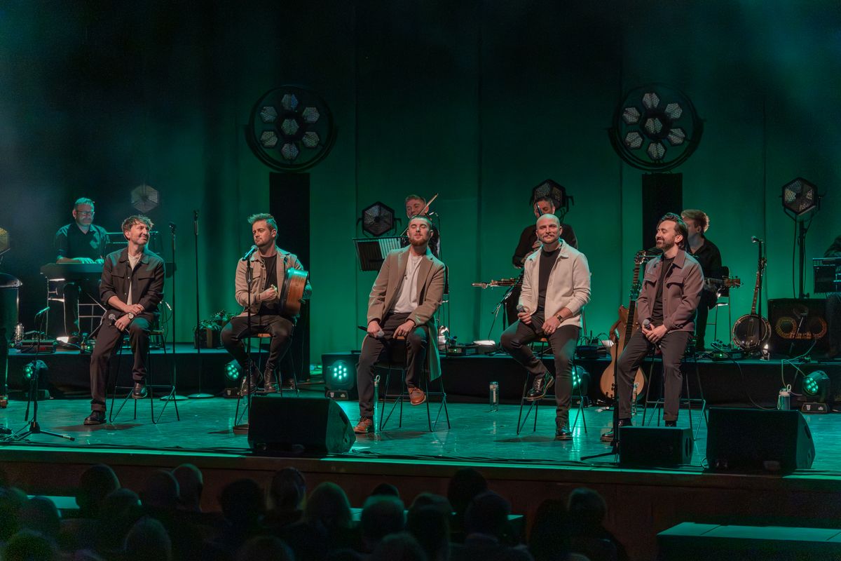 Five band members sat on stools on stage with a green light background 