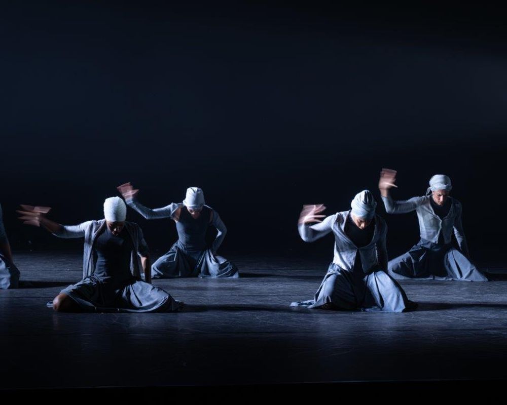 Group of dancers on stage wearing blue and grey outfits, a light casts a shadow over them