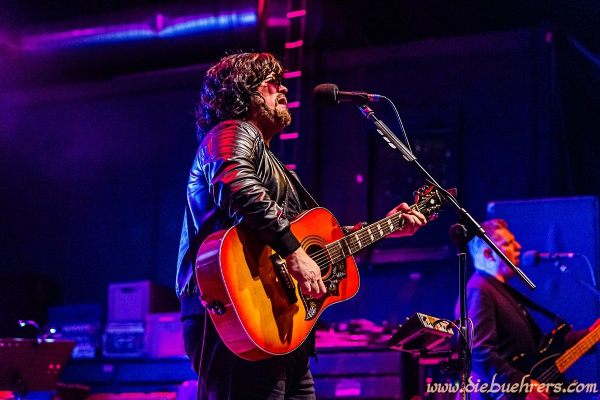 A musician with curly hair and sunglasses plays an acoustic guitar on stage. He wears a black leather jacket and sings into a microphone, creating a vibrant, energetic atmosphere with blue and purple stage lighting in the background.