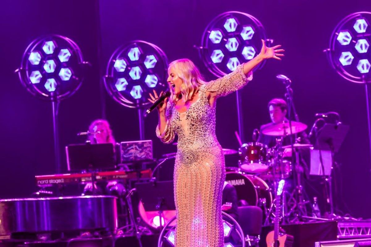 Singer in a sparkly dress performs passionately on stage, holding a microphone. Bright purple lights and a band with drums and keyboard in the background.
