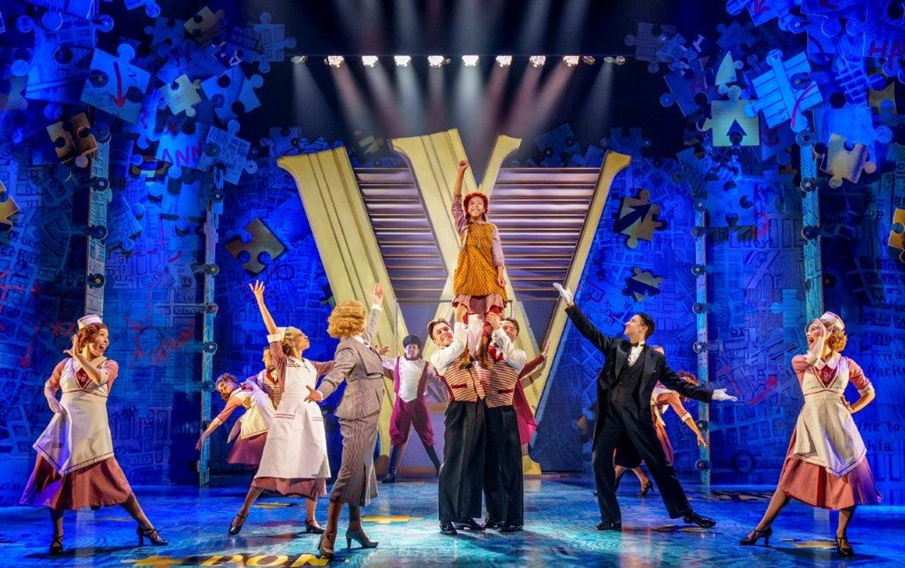 Stage performance with enthusiastic actors in colorful 1940s-style costumes. A girl in orange is lifted high, surrounded by a lively ensemble, under bright lights.