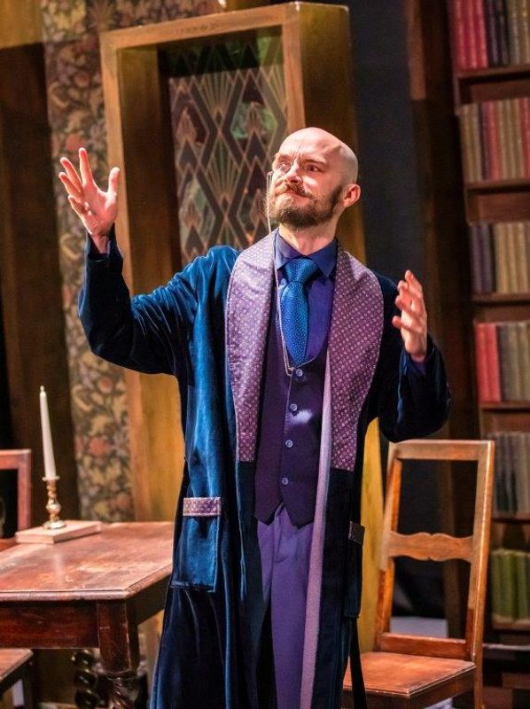 A man in a blue robe gestures expressively on a stage set with a wooden table, chairs, and bookshelves. The scene has a dramatic and theatrical atmosphere.