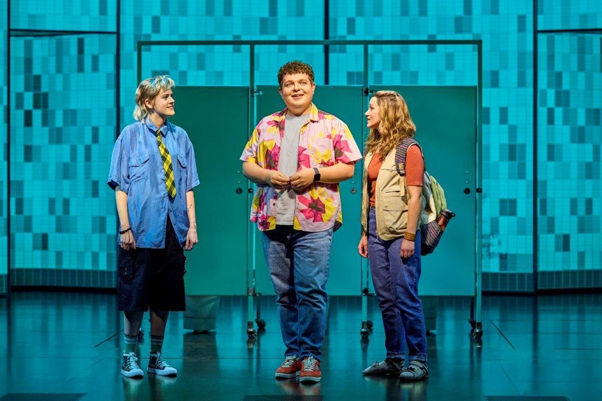 Three actors stand on stage in front of teal bathroom stalls. They wear casual, colorful clothing, conveying a light, humorous tone.