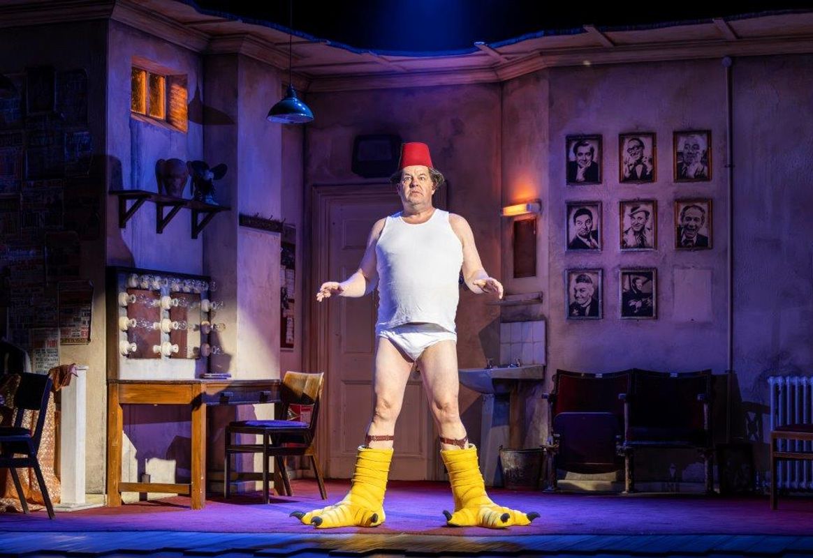A man in a comedic outfit, wearing a fez, big yellow boots and undershirt, sits onstage with an exaggerated expression. The room is cluttered and lively, featuring a vintage vanity with lights, posters, and props. The atmosphere is theatrical and whimsical, evoking humor and nostalgia.
