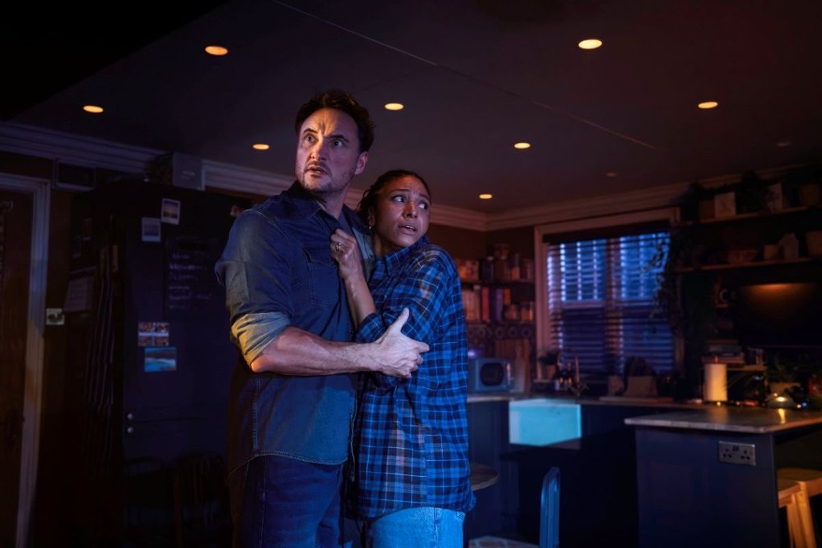 Two people stand close together in a dimly lit kitchen, bathed in blue light, holding each other and looking anxiously off to the side as if reacting to something unseen. The space feels tense and eerie, with everyday kitchen items visible in the background.