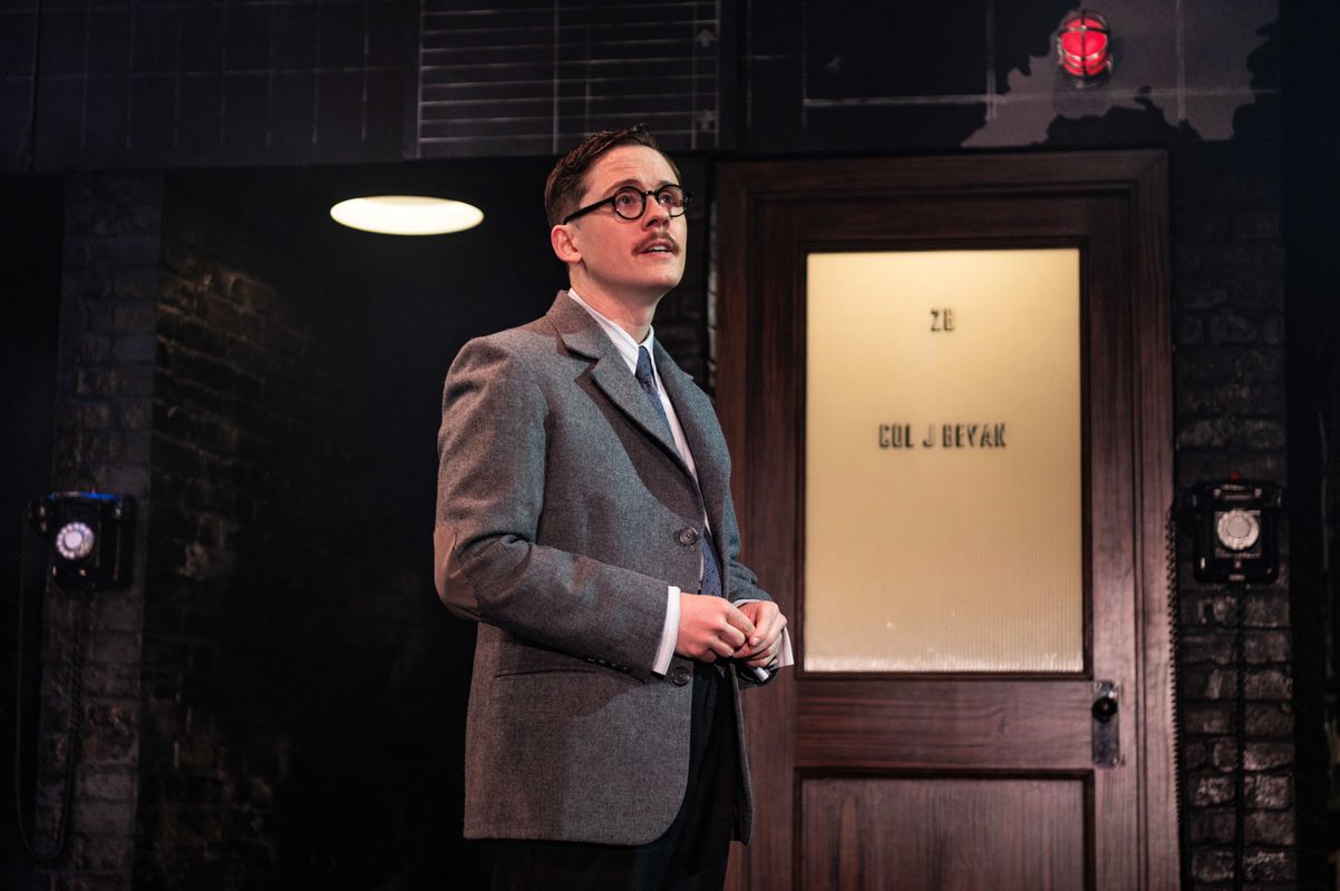 A man in a vintage suit stands on stage, looking up thoughtfully. The background features a wooden door with frosted glass and dim stage lighting.