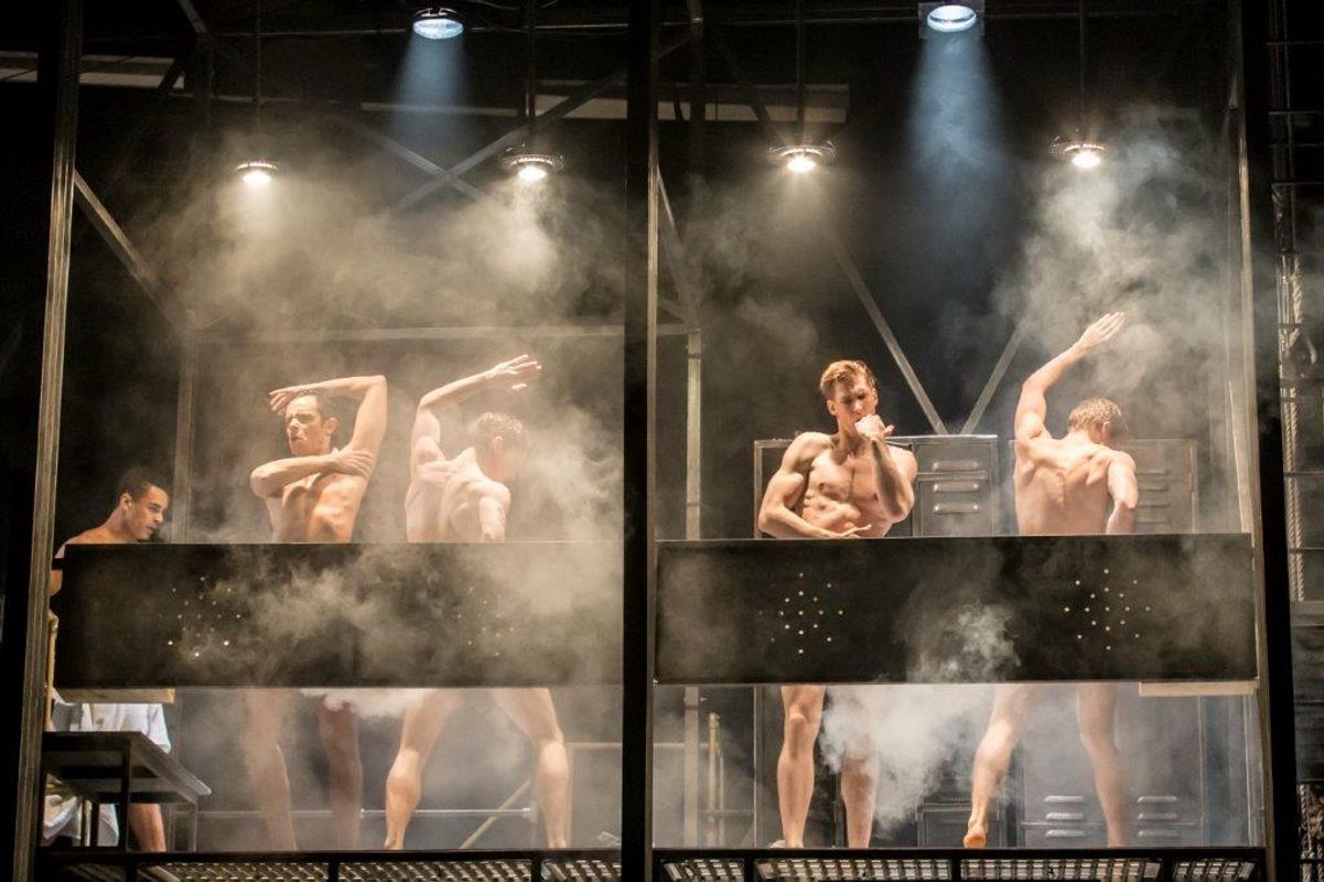 Four men showering on stage, surrounded by mist and dramatic lighting. They stand behind metal fixtures, creating an intense and artistic atmosphere.