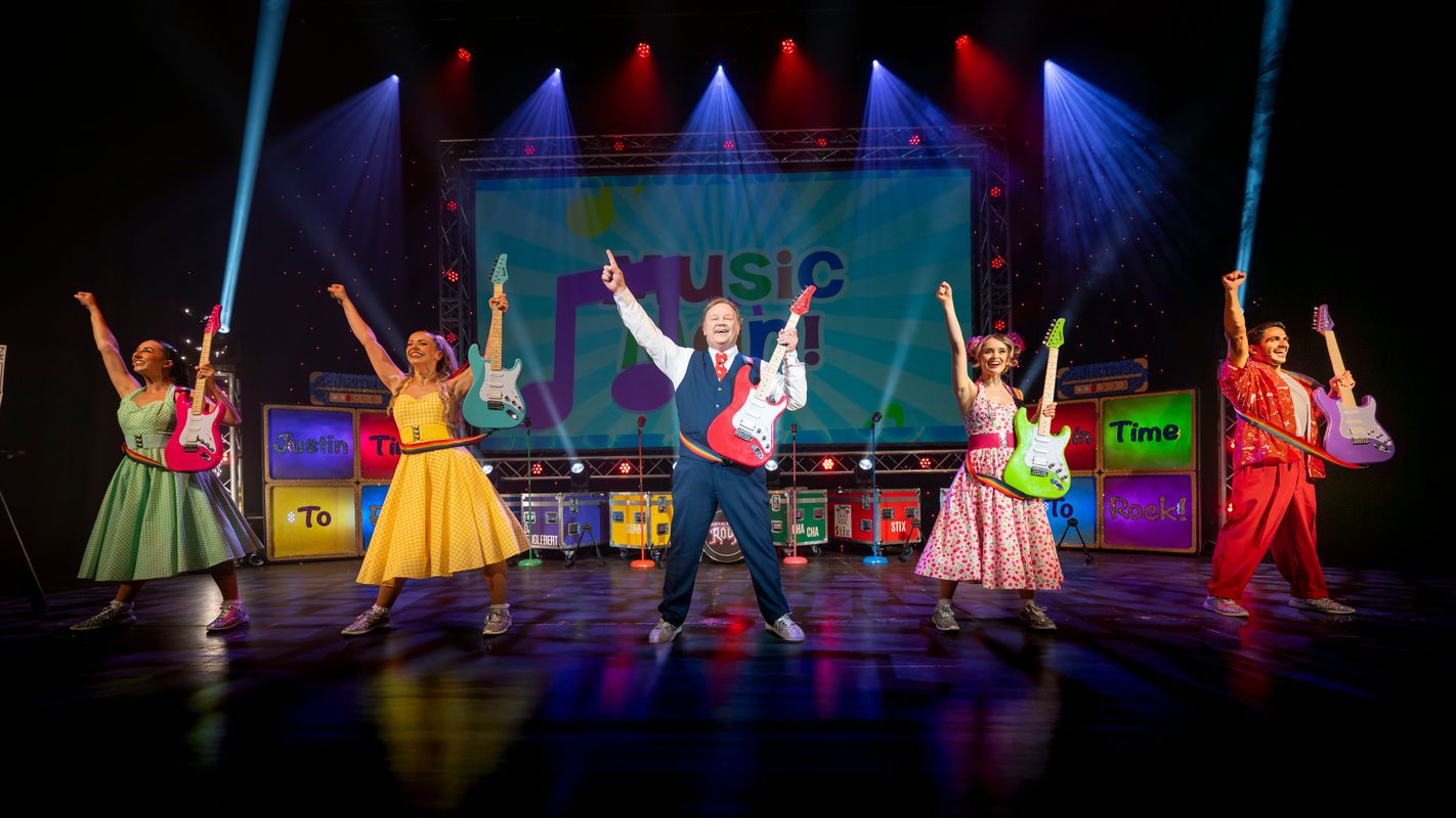 Justin and cast members hold their right hand in the air and hold an electric guitar in their left  hand.