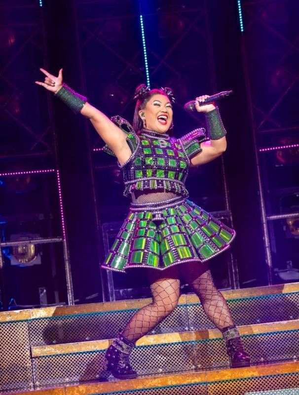 A person wearing a green and black checked outfit stands with their fingers raised in the air.