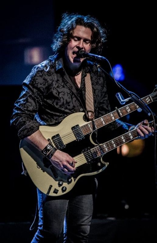 A person is performing on stage, playing a double-neck Gibson guitar and singing into a microphone. The setting is dimly lit, creating a dramatic atmosphere.