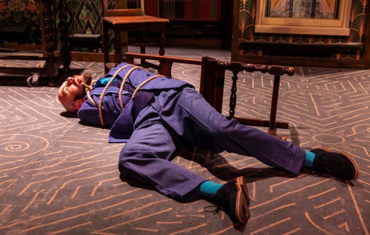 A man in a blue suit lies on a patterned floor, bound with rope, next to a fallen chair. The scene conveys tension and drama.
