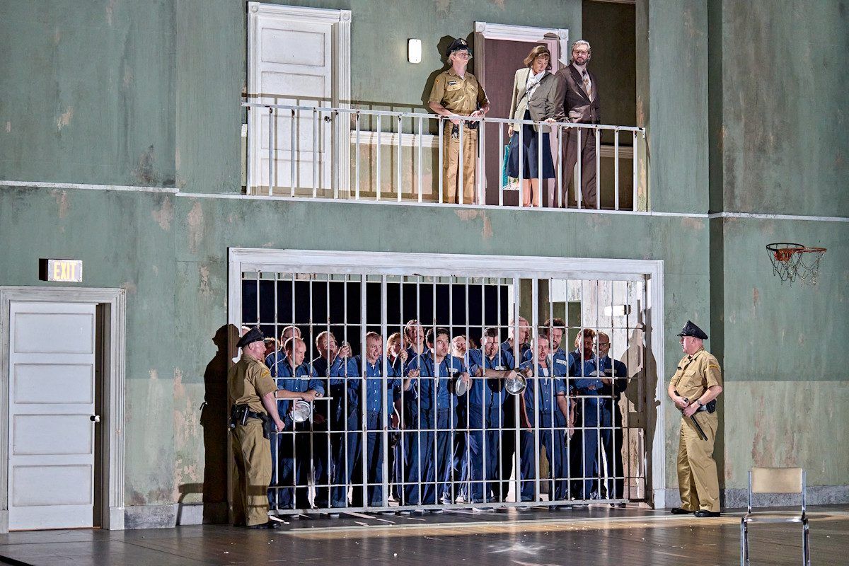 There are a group of inmates behind a cell door and there are two guards either side and people stood above the inmates