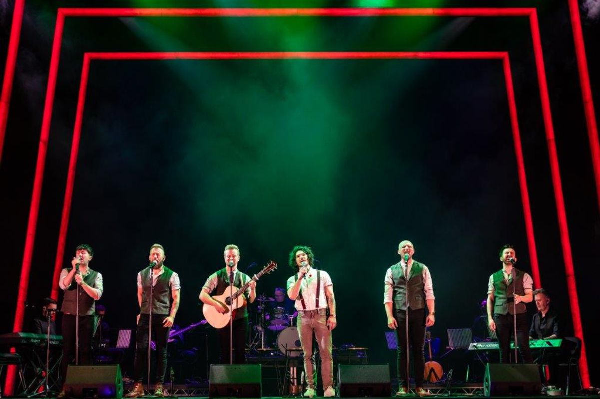 Six male performers on stage, singing with microphones. One holds a guitar. They're wearing vests under dramatic red and green lighting, creating an energetic atmosphere.