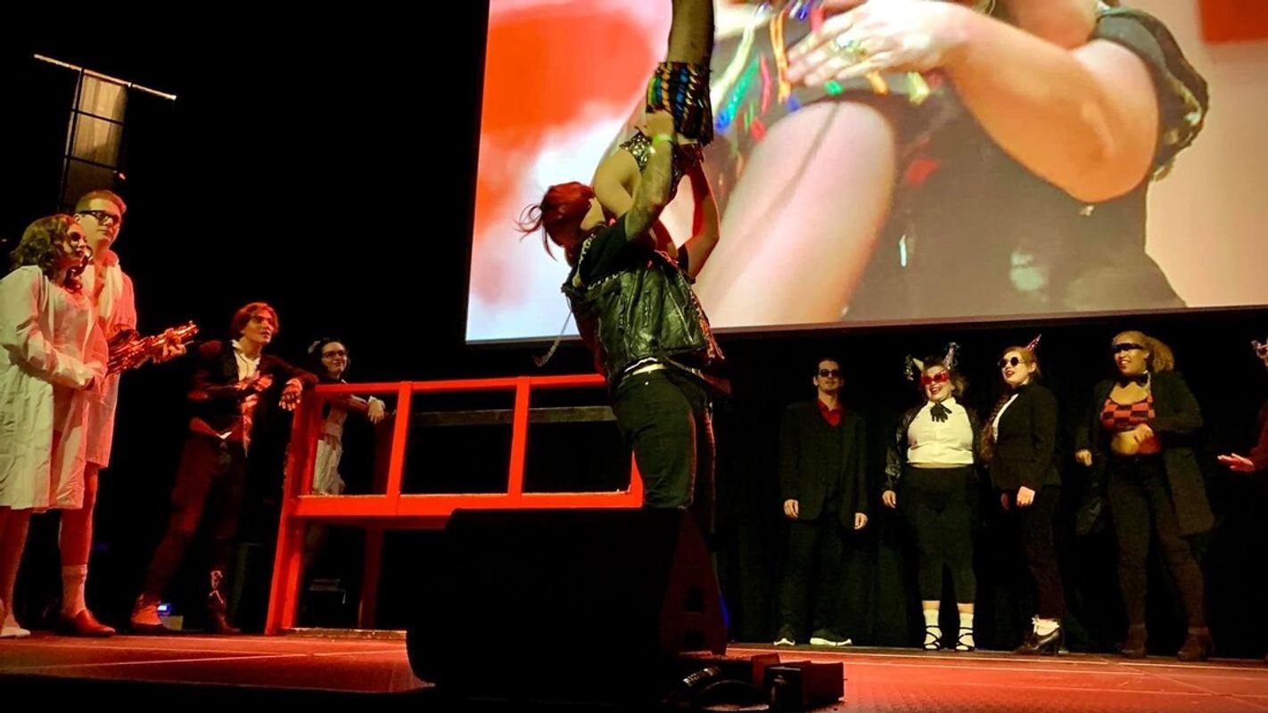 Group of people on stage. Those stood at the back are wearing black suits, white shirts and party headwear. Two people to the left are wearing lab coats. The two characters in the middle are dancing, one is holding the other one up in the air.