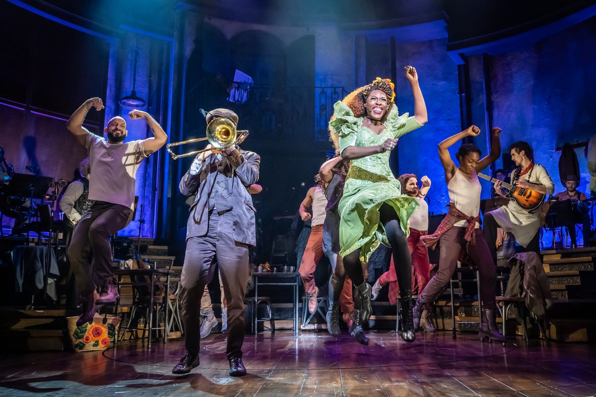 An actor in a green dress jumps in the air with a trumpeter playing next to them and ensemble members dancing behind. 