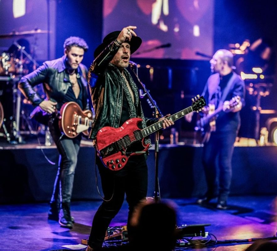 A band performs energetically on stage with vibrant lighting. The person in the foreground plays a red electric guitar and interacts with the audience. A guitarist in the background plays alongside another musician. Drums and additional instruments are visible behind them.