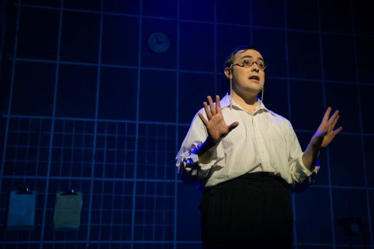 A person stands on stage under blue lighting, wearing glasses and a white shirt, with hands raised expressively. A clock and clipboards are visible on the grid-patterned wall behind, suggesting a dramatic or theatrical setting.
