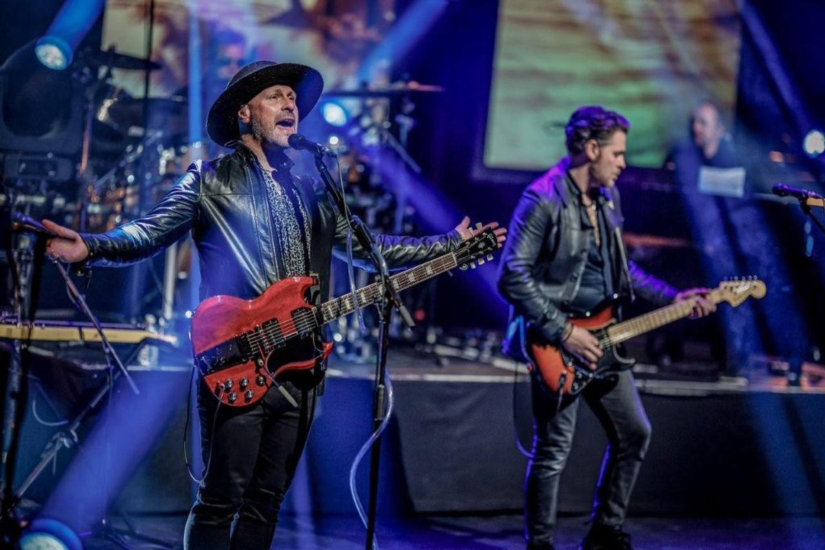 A band performs energetically on stage. The lead singer, wearing a hat, plays a red electric guitar. Another member plays a different electric guitar. The stage is lit with colorful lights and a drummer is visible in the background.