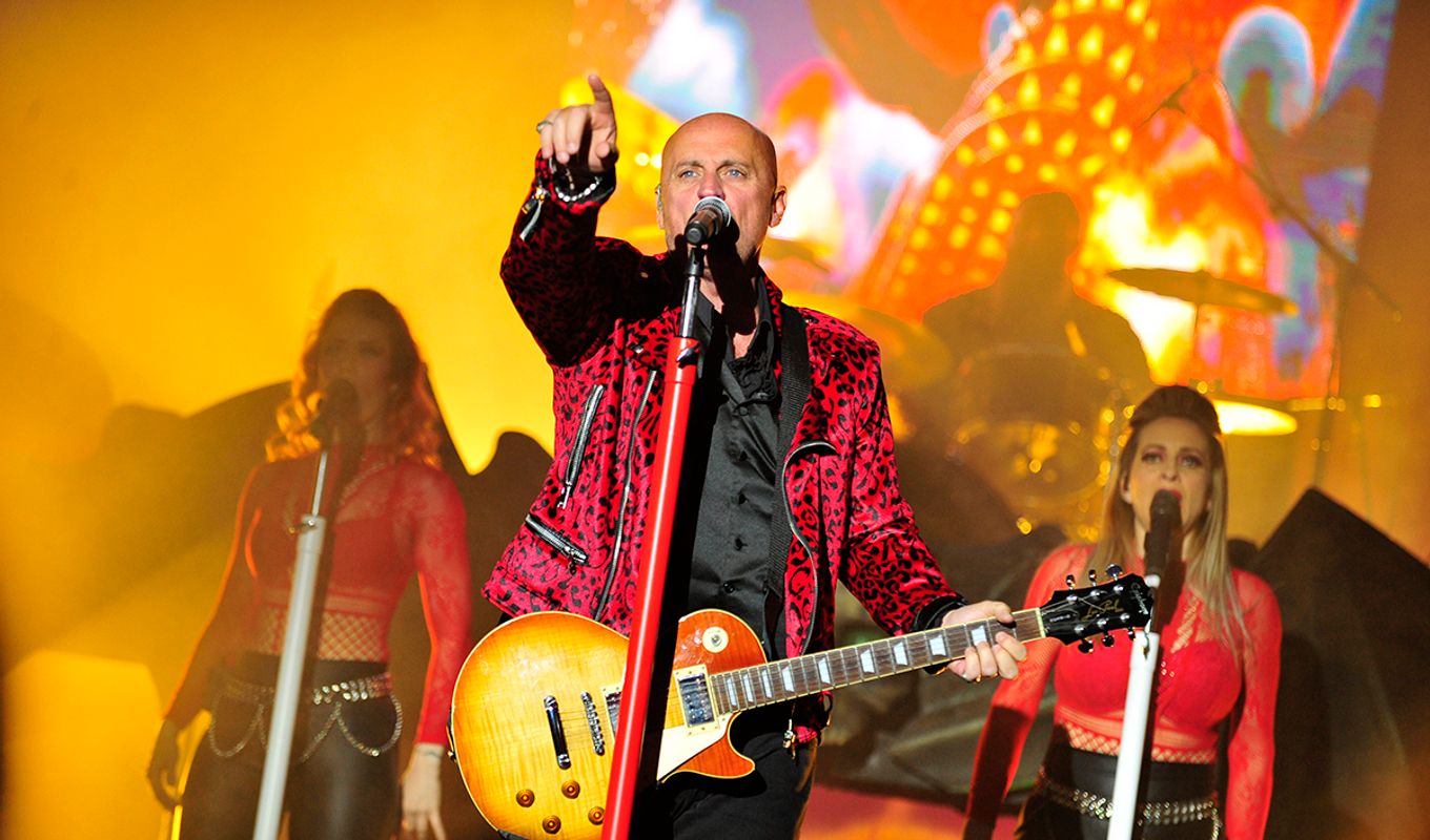 Steve Steinman in a red leopard jacket passionately performs on stage, pointing while holding a guitar. Backing vocalists in red and a drummer enhance the vibrant scene.