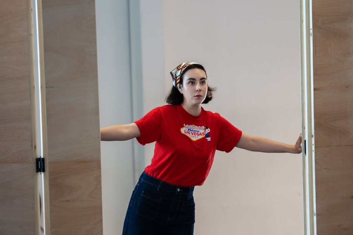 A woman in a red t-shirt and bandana is stood in a doorway looking concerned