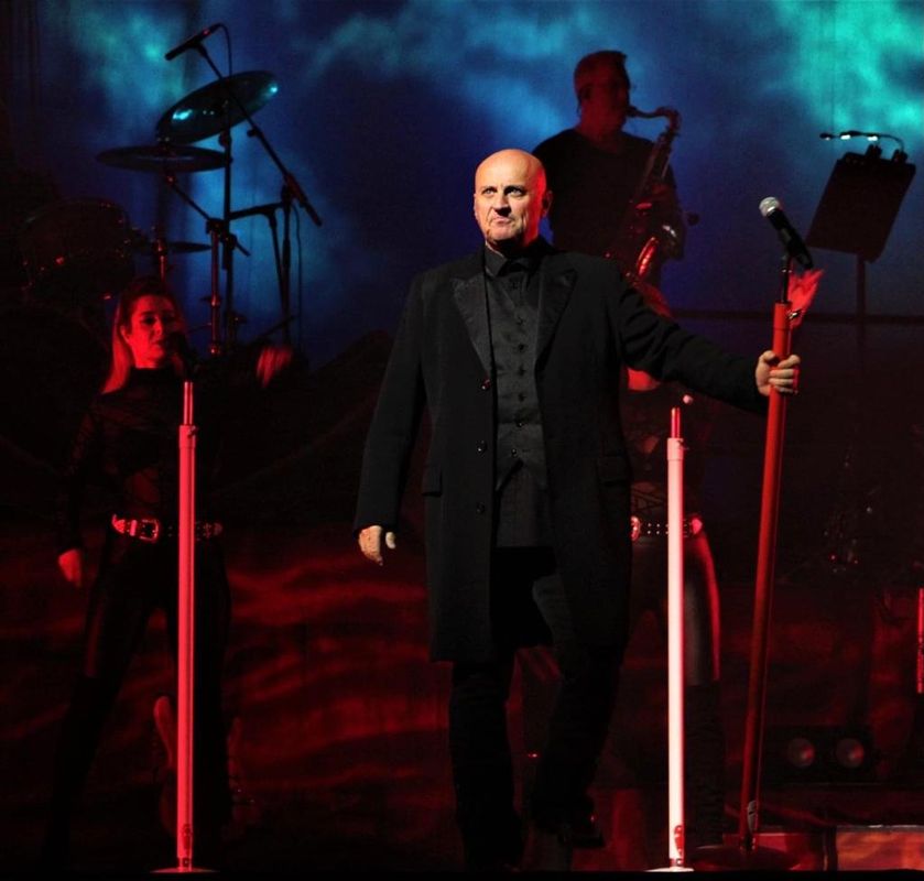 Steve Steinman stands confidently on stage holding a microphone stand, wearing a dark coat. Dim red lighting, blue background, and backup musicians create a dramatic atmosphere.