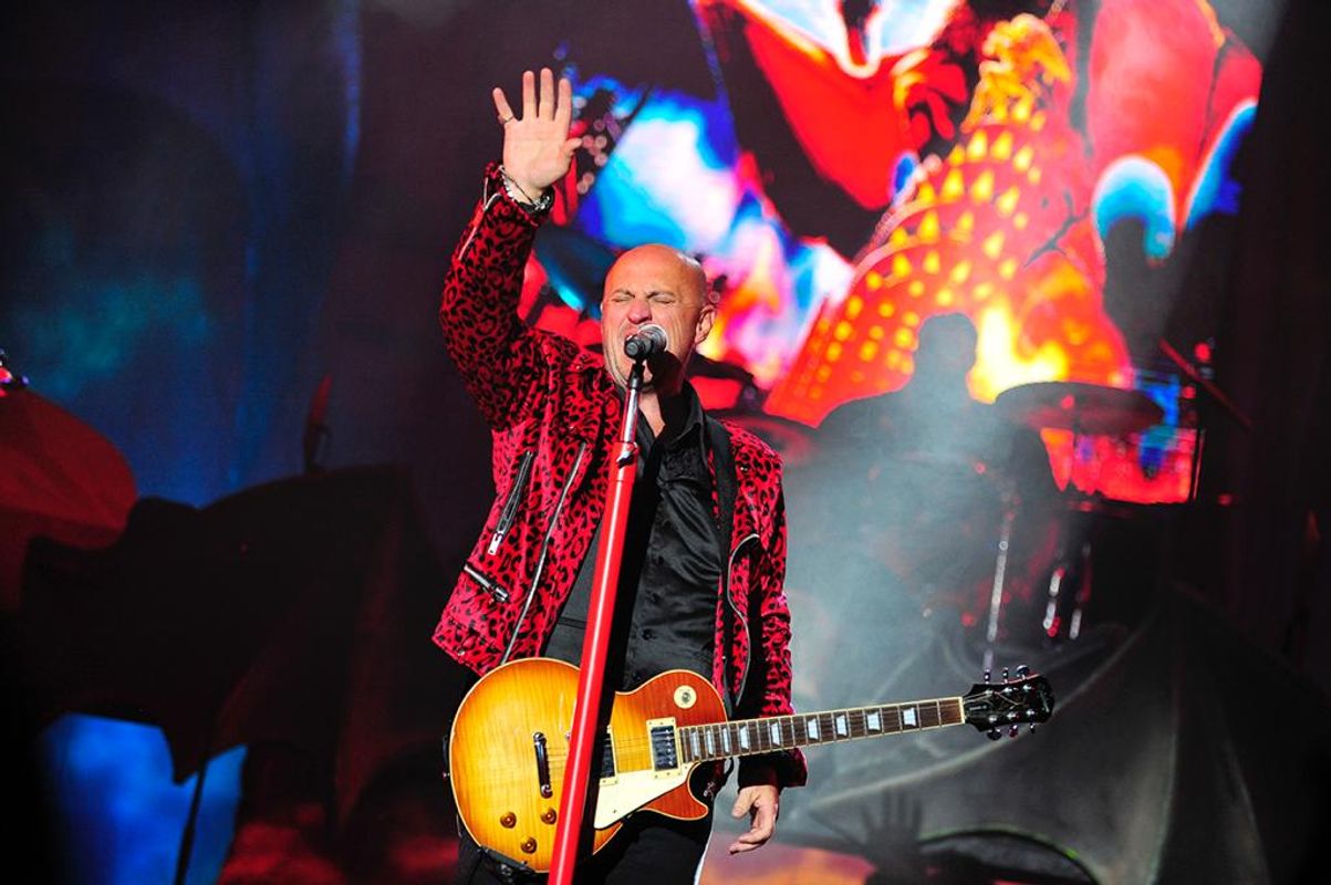 Steve Steinman on stage wearing a red leopard print jacket, holding a guitar and raising his hand. Vibrant abstract background and dynamic lighting. Energetic atmosphere.