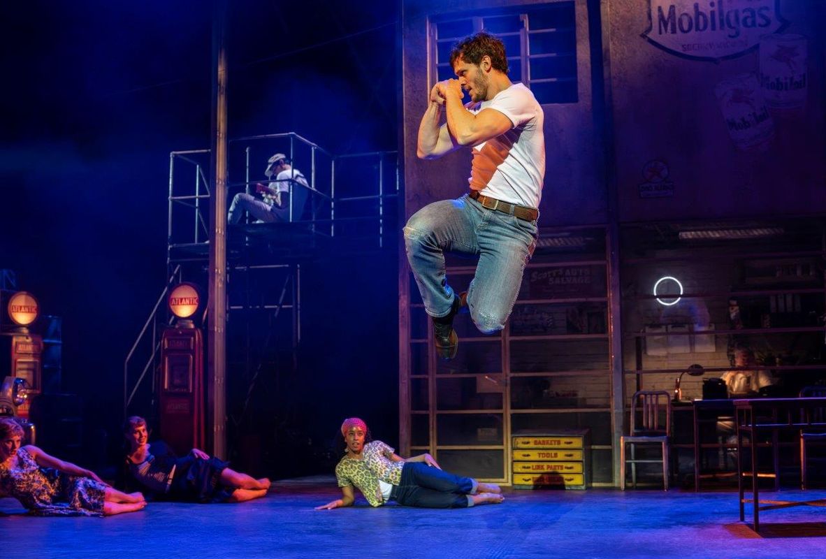 Performer energetically leaps on stage in a dynamic dance scene, set against an urban backdrop with graffiti, exuding excitement and passion.