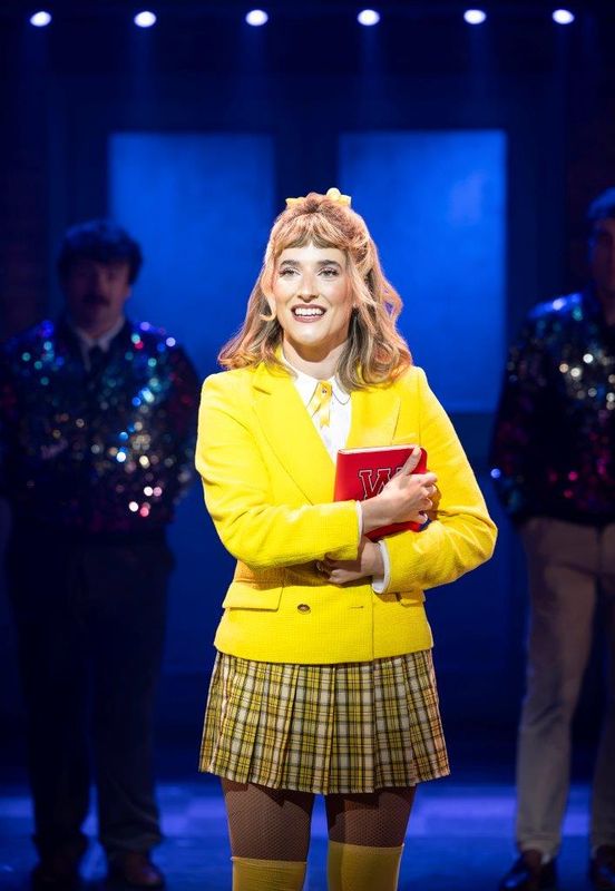 A girl in a yellow blazer looks on and is smiling