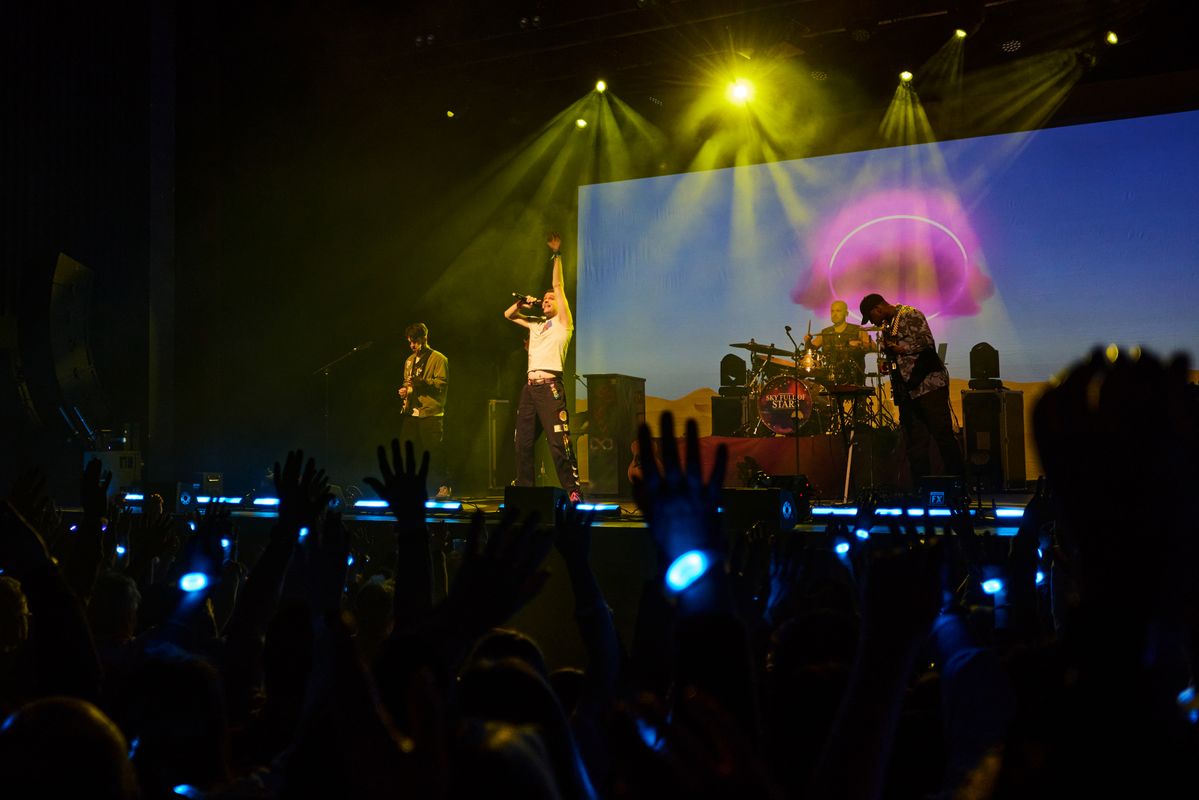 A band performs energetically on stage with vivid yellow and purple lights. A crowd with raised hands and glowing wristbands creates an exhilarating atmosphere.