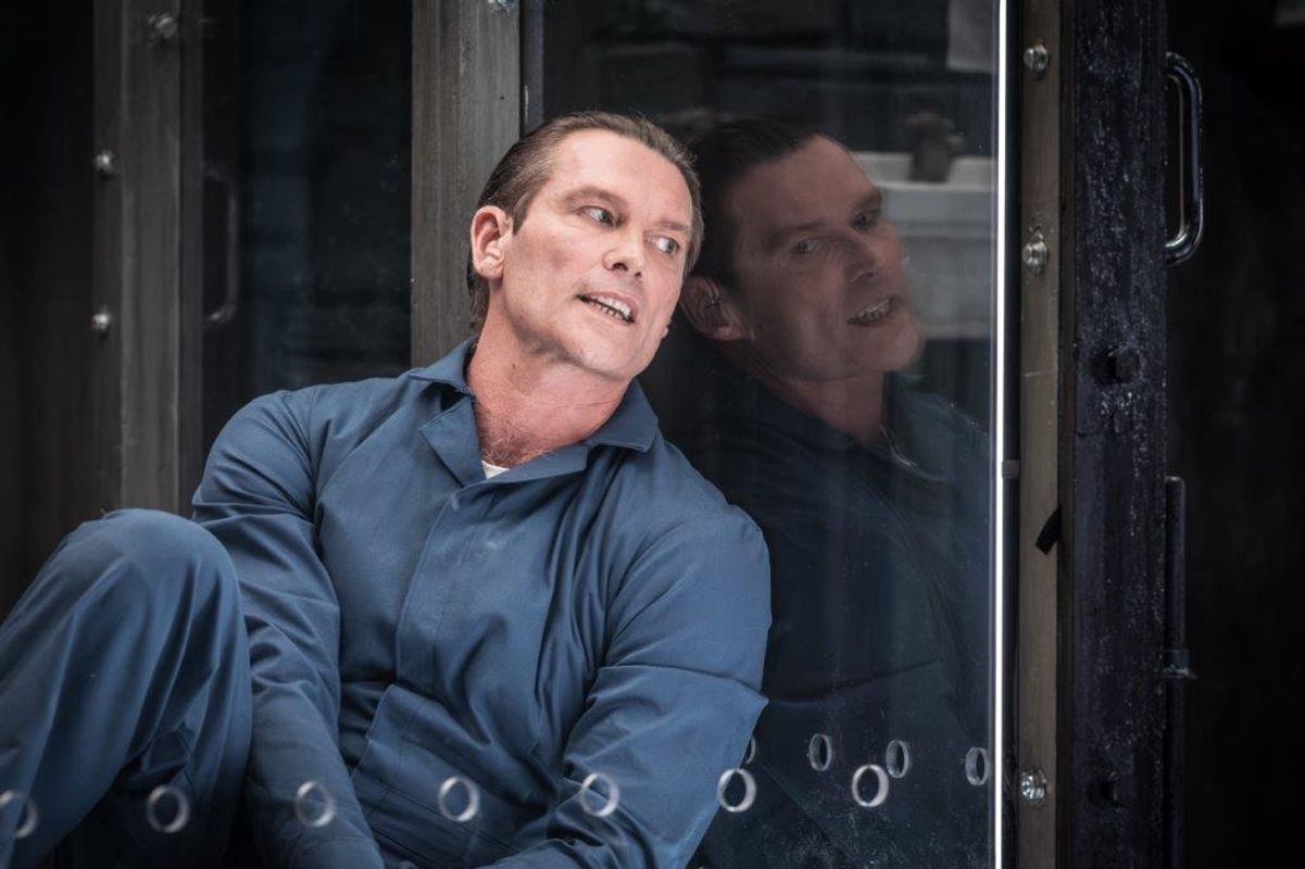 A man wearing navy overalls is sat in a glass cell.