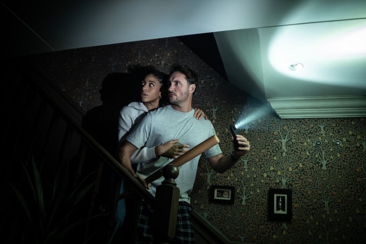 A man in pajamas holds a flashlight and a bat, looking anxious, with a woman clutching his arm.