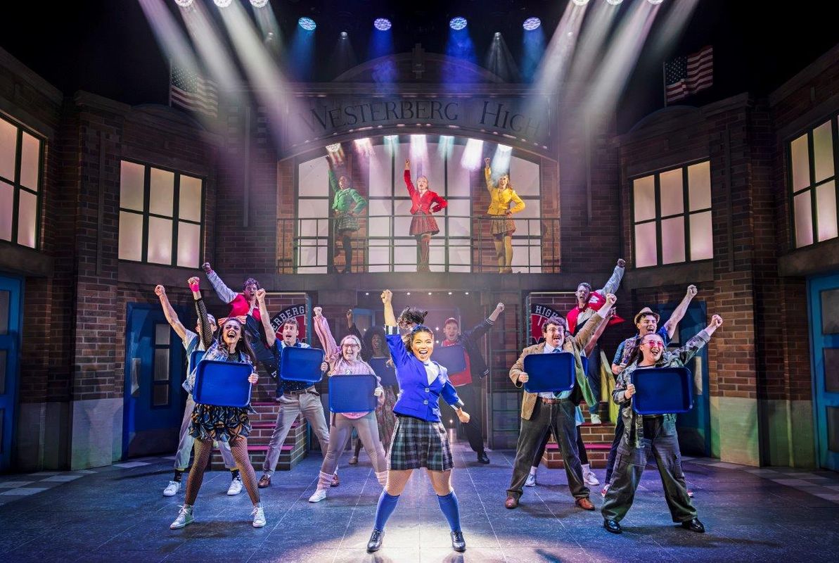 A vibrant stage scene shows performers in school uniforms celebrating under colorful lights. The backdrop reads "Westerberg High." Some hold blue trays aloft energetically.
