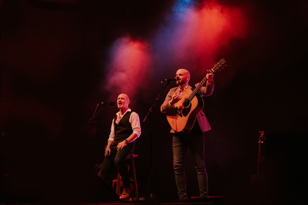 One person is singing into a microphone and the other person singing into a microphone and playing the guitar